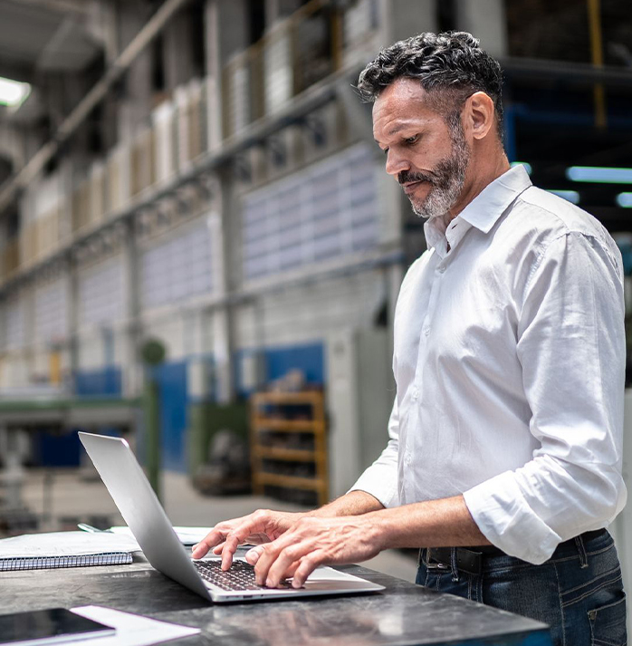 Man in a white shirt working on a laptop in an industrial factory setting.