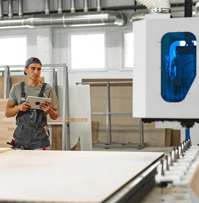 Worker in a cap and overalls using a tablet to monitor a machine in a furniture manufacturing facility.